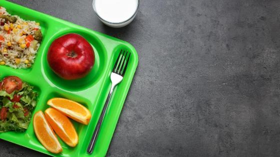 Gesunder Schulmittagessen mit reis, Gemüse, frischem Obst liegt auf einem grünen Tablett in einer Schul-Mensa.