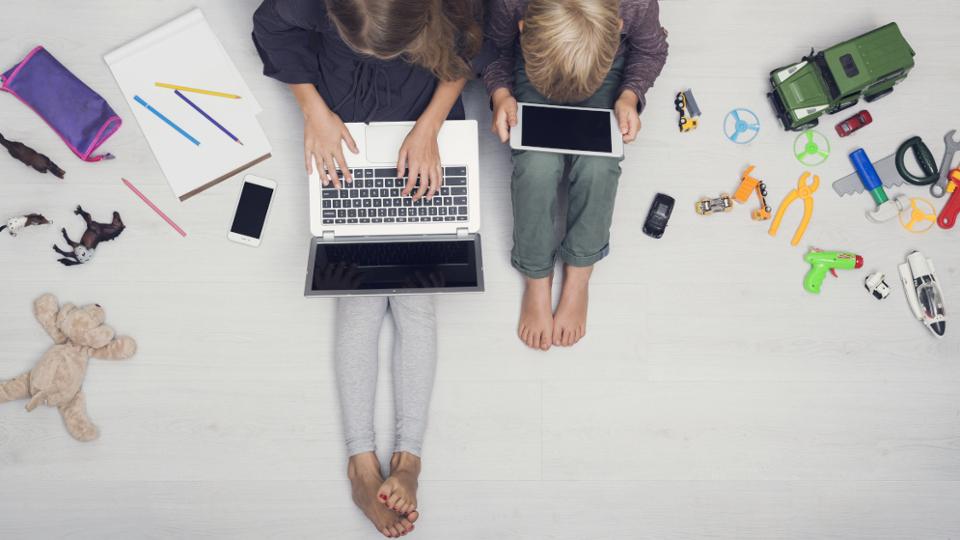 Zwei Kinder sitzen auf dem Boden und schauen in ein Tablet und ein Notebook. Spielsachen liegen daneben. Von oben fotografiert