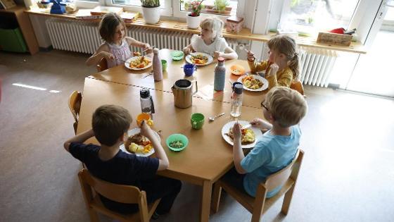 Fünf Kinder sitzen in einer Kita an einem Holztisch und essen ihr Mittagessen.