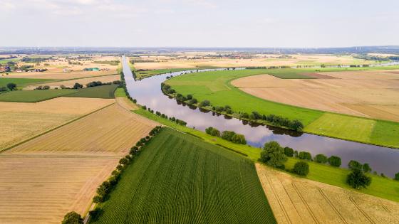 Luftaufnahme von Feldern, durch die ein Fluss führt, von dem ein Kanal abgeht. im Hintergrund eine Biogasanlage